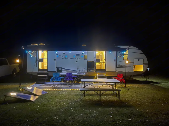 An inviting RV rental campsite at night, featuring a brightly lit RV, outdoor seating, a picnic table, and cornhole boards.. Forest River Salem Hemisphere HL 2021