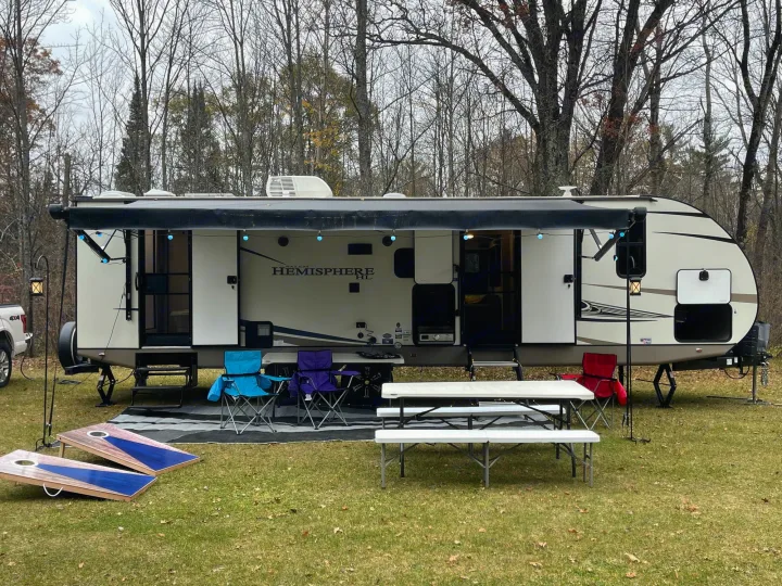 An rv rental set up at a campsite with a travel trailer, extended awning, blue lights, chairs, tables, and cornhole.. Forest River Salem Hemisphere HL 2021