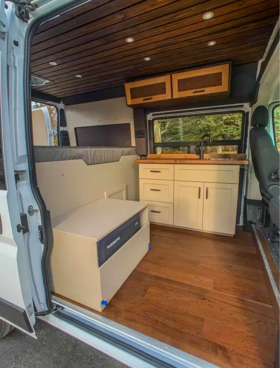 Modern RV rental camper van interior. Features wood plank ceiling, full kitchen, bed area, and Dometic fridge on wood floors.. Dodge Ram 1500 2021
