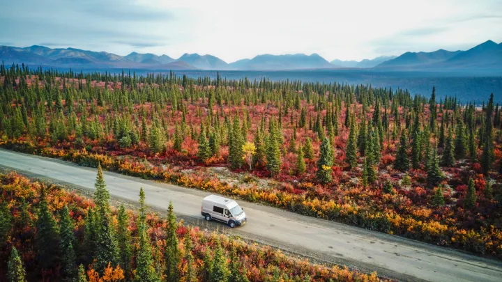 An RV rental travels through a vibrant fall forest, rich with green, red, and orange foliage, against distant mountains. Ideal road trip.. Dodge Ram 1500 2021
