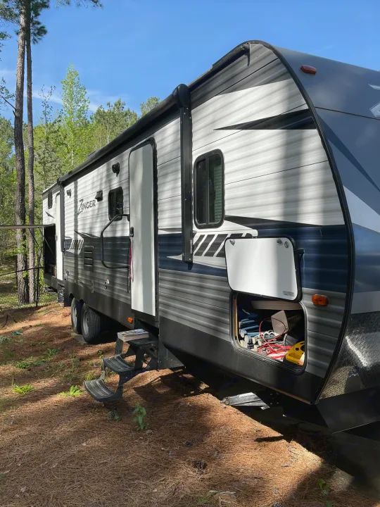 RV rental parked at a serene campsite, showing open storage and entry steps. Ideal for outdoor adventures in nature.. Crossroads RV Zinger 2019