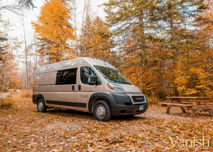 Autumn adventure: A modern tan RV rental, a converted camper van, is nestled in a colorful fall forest campsite with a picnic table.. Ram Promaster 2022