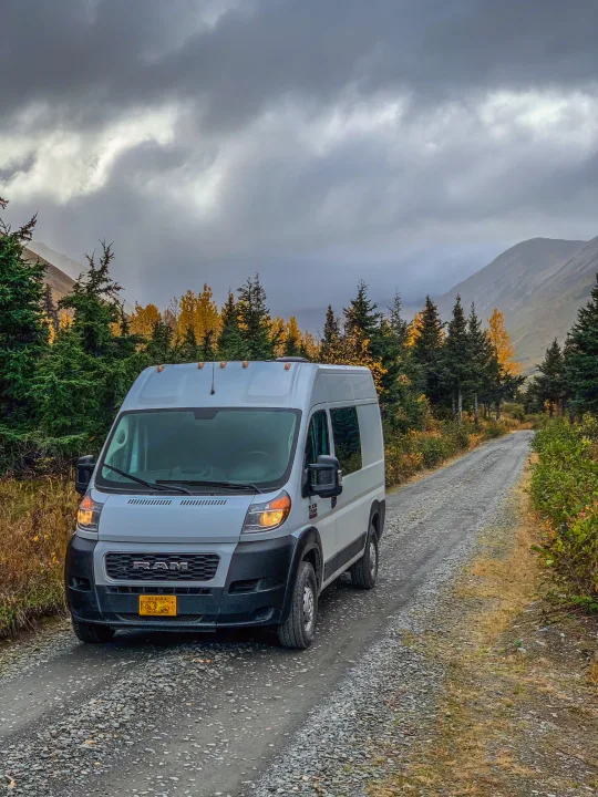 White RV rental van on a gravel road in the Alaskan mountains, surrounded by vibrant autumn trees under a cloudy sky.. Dodge Ram 1500 2021