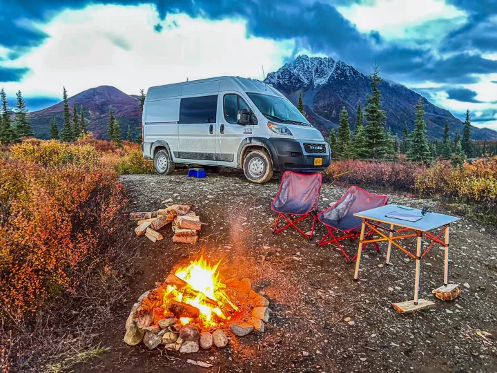 RV rental adventure: a white van camp setup with a warm campfire, chairs, table, and stunning mountain backdrop.. Dodge Ram 1500 2021