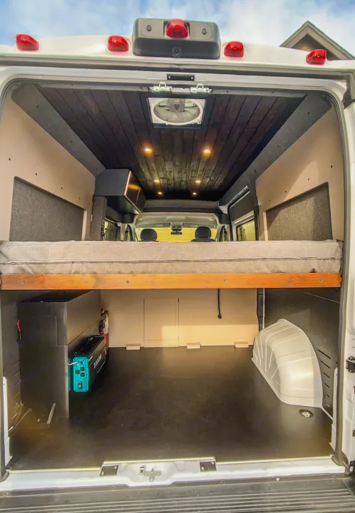 Inside this RV rental van conversion: a cozy elevated bed, sleek dark wood ceiling, and extensive lower storage with power system.. Dodge Ram 1500 2021