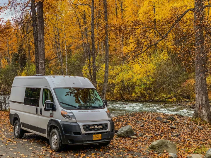White RV rental parked by a vibrant autumn forest and river. Perfect for scenic fall adventures.. Dodge Ram 1500 2021