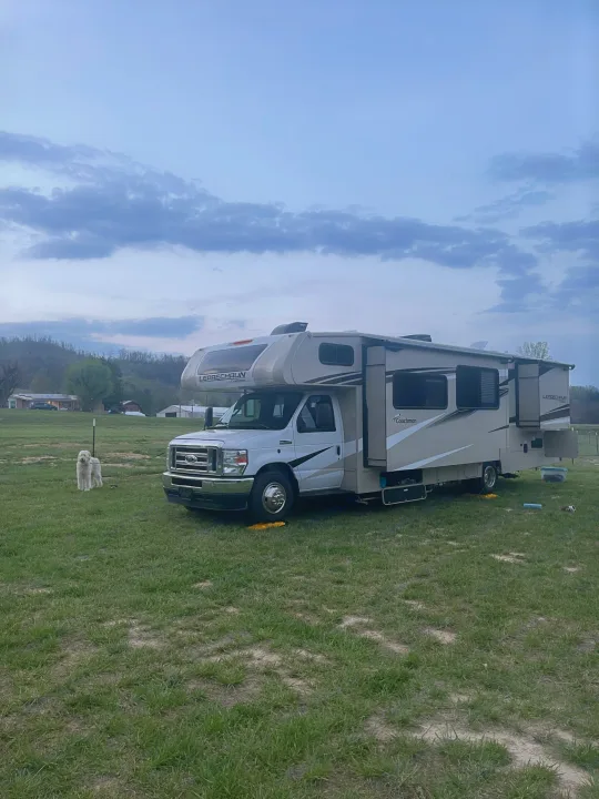 This RV rental, a Coachmen Leprechaun motorhome, is parked in a grassy field at dusk with a fluffy white dog nearby.. Coachmen Leprechaun 2023