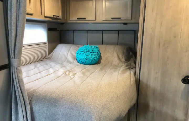 Cozy RV rental bedroom featuring a light-colored bed, teal decorative pillow, and wooden overhead cabinets.. Forest River Coachmen Cross Trail XL 2022