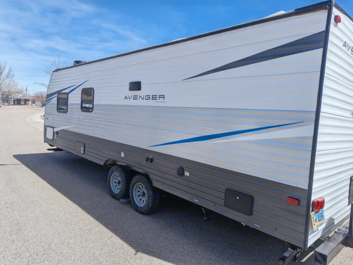 Side view of a white and gray Avenger travel trailer with blue accents, ideal for your next RV rental trip, parked on asphalt.. Avenger 26bk 2021