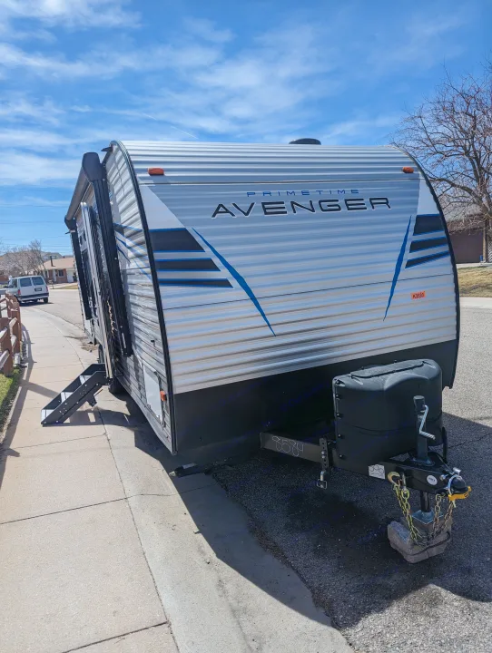 A Prime Time Avenger travel trailer, ideal for an RV rental, parked on a street. Its front and side with steps are visible under a blue sky.. Avenger 26bk 2021