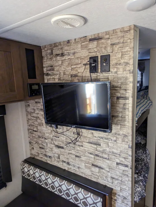 RV rental interior: A mounted TV on a faux stone wall, with an entertainment system, seating, and bunk beds visible.. Avenger 26bk 2021