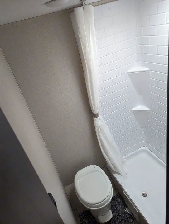 Inside a compact RV rental bathroom: features a modern toilet, standing shower with curtain, & built-in shelves.. Avenger 26bk 2021