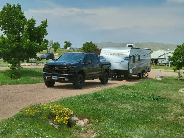 Black Chevy truck towing a silver Prime Time Avenger travel trailer (RV rental) in an RV park.. Avenger 26bk 2021