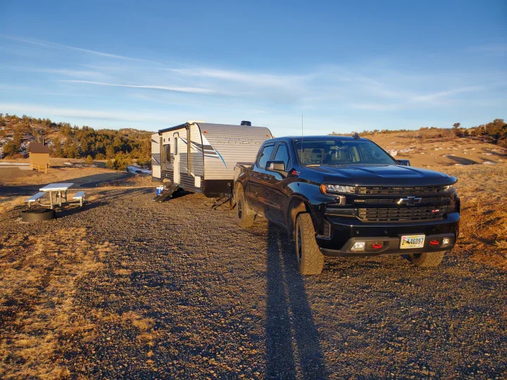 RV rental adventure: Black truck towing a travel trailer at a sunny gravel campsite with a picnic table & mountains beyond.. Avenger 26bk 2021