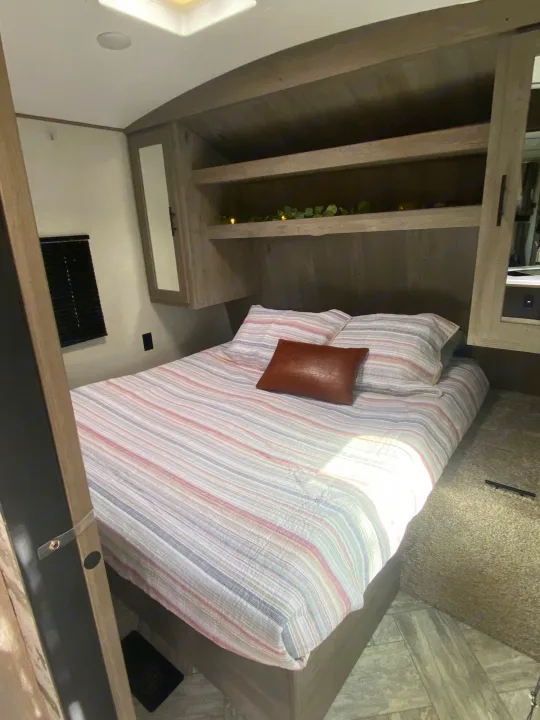 Comfortable RV rental bedroom featuring a striped bed, brown accent pillow, built-in shelving, and a window. Ideal for travelers.. Keystone Springdale 2021