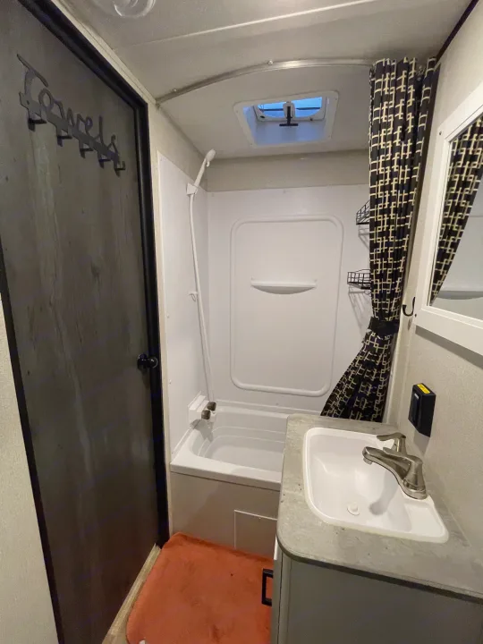 An RV rental bathroom, showcasing a compact tub/shower, sink, and dark door with a "TOWELS" hanger.. Keystone RV Hideout 2022