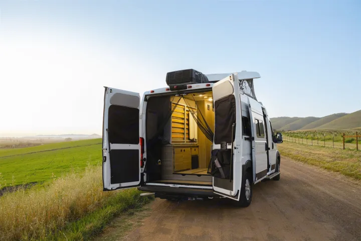 A white RV rental camper van with open rear doors on a dirt road, showcasing its inviting interior space & hill views.. Winnebago Solis 2022