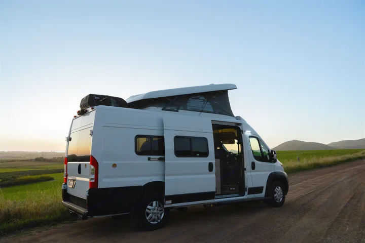 This white pop-top camper van, ideal for RV rental adventures, is parked on a dirt road amidst green fields at sunset.. Winnebago Solis 2022