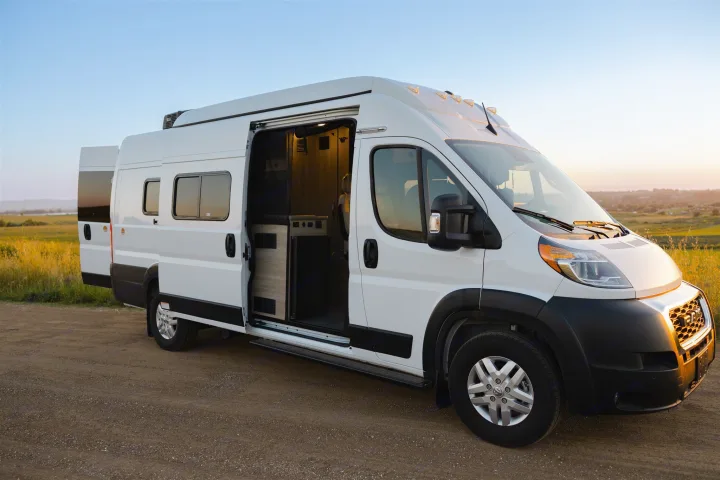 Discover adventure with this white camper van RV rental. Parked on a dirt road, its open doors reveal a cozy interior, bathed in sunset light.. Winnebago Solis 2022
