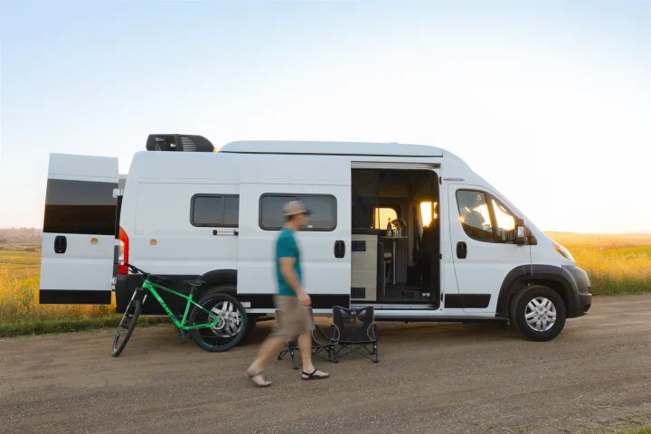 Your RV rental adventure awaits! A camper van with open doors, bike, chairs, and a person on a dirt road at sunset.. Winnebago Solis 2022