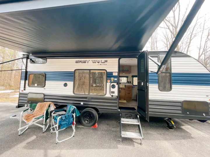 An inviting Grey Wolf rv rental travel trailer at a campsite, with its awning extended and two chairs set up for relaxation.. Cherokee MKSE22 2023