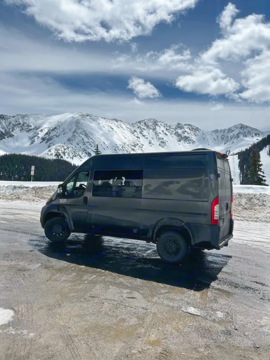 Explore winter peaks with this RV rental! Dark camper van parked on a snowy, muddy road under cloudy mountain skies.. Ram Promaster 2022