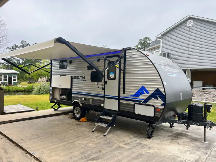 This travel trailer RV rental boasts an extended awning with blue lights, steps, and outdoor kitchen, by the water.. Forest River Coachmen Catalina Summit 2022