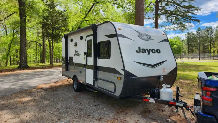 A white & grey Jayco travel trailer, an RV rental, parked on gravel beside a road, surrounded by vibrant green trees.. Jayco Jay Flight 2022