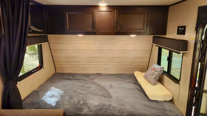 Inside a comfortable RV rental bedroom, featuring a large bed, overhead cabinets, and side windows offering scenic views.. Jayco Jay Flight 2022