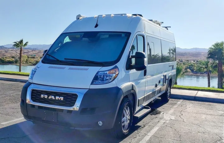 White Ram ProMaster RV rental van parked by a scenic lake. Features include large windows and roof racks under a sunny sky.. Thor Sequence 2022