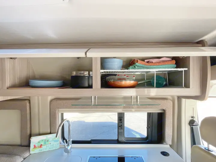 RV rental kitchen: Open overhead cabinets with dishes, pan, and towels. Countertop has a sink, faucet, and small stovetop.. Thor Sequence 2022