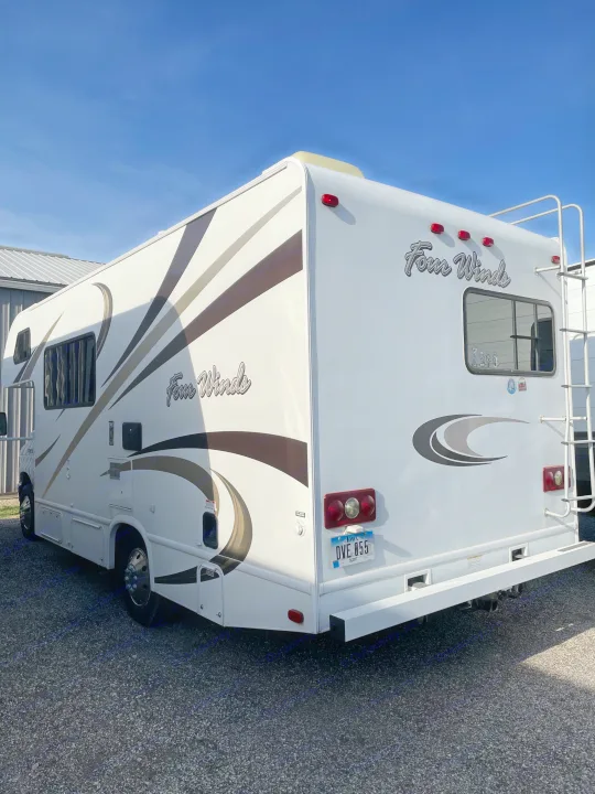White Four Winds motorhome RV rental, with brown/grey graphics & rear ladder, parked on gravel under a blue sky.. Thor Four Winds 2015