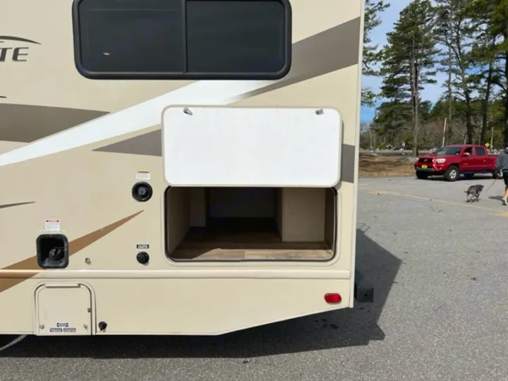 Rear side of an RV rental, showing an open exterior storage bay and utility hookups. Parked with a truck and dog nearby.. Thor Motor Coach Freedom Elite 2017