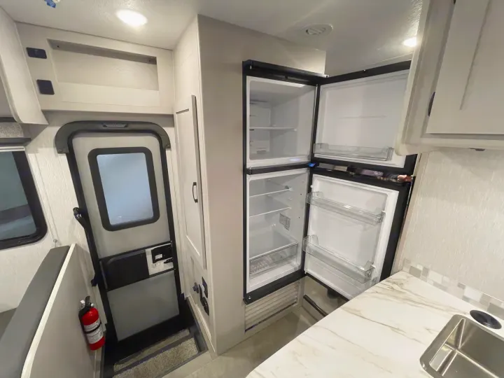 Inside an RV rental: Open refrigerator, kitchen counter with sink, entry door, dinette, and overhead storage. Modern and functional.. Coachmen Freelander 2024