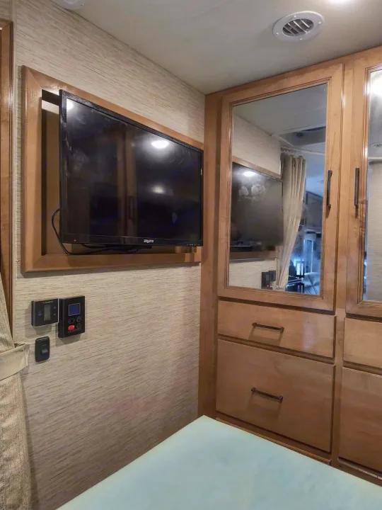 Inside an rv rental: a wall-mounted TV, Dometic controls, and mirrored cabinets with storage drawers beside the bed.. Thor Quantum 2021