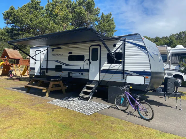 A travel trailer rv rental set up at a campground with an awning, outdoor rug, picnic table, and bicycle.. Keystone Springdale 2021