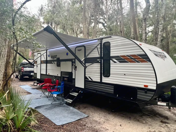 An RV rental and truck are set up at a rustic campsite, with an extended awning, lights, and outdoor chairs.. Forest River Wildwood X-Lite 2022