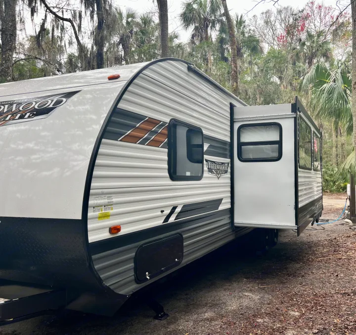 White Forest River Wildwood X-Lite rv rental at a shaded campsite with its slide-out extended, surrounded by trees.. Forest River Wildwood X-Lite 2022