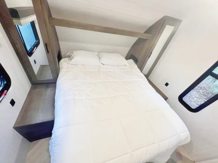 Inside an RV rental: a cozy bedroom with a white bed, built-in shelving, and windows. Ideal for travel.. Forest River Wildwood X-Lite 2022