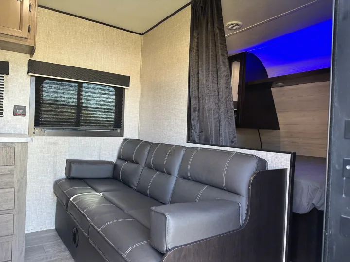 Modern RV rental living space featuring a black sofa, window blinds, and a separate bedroom with blue lighting.. Jayco Jay Flight SLX 2022