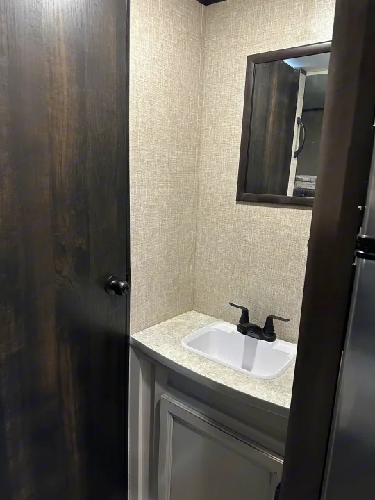 Inside an RV rental: a cozy bathroom vanity with a white sink, black faucet, mirror, and dark wood door.. Jayco Jay Flight SLX 2022