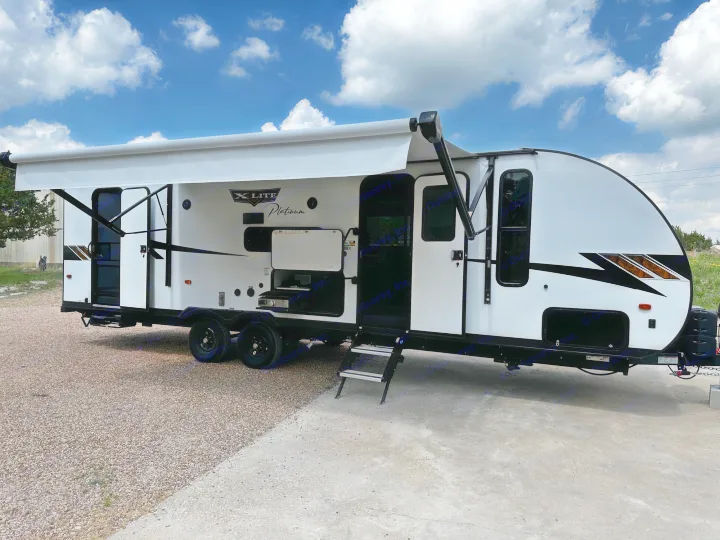 Side view of a white & black travel trailer with an extended awning, parked on gravel & concrete. Great for an RV rental.. Forest River Wildwood X-Lite 2023