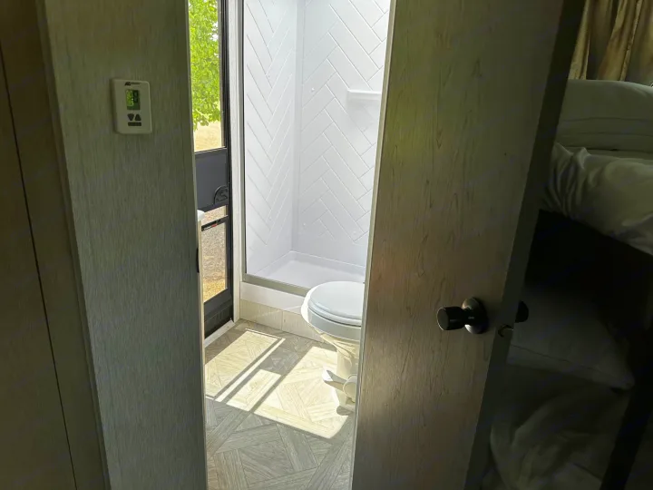Inside this RV rental: A well-lit bathroom with shower, toilet, and thermostat. Bunk beds visible. Sunlit floor.. Forest River Wildwood X-Lite 2023