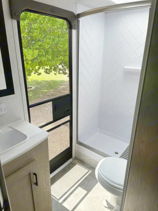Inside an RV rental bathroom: modern shower, toilet, and sink, with a clear view of green trees outside.. Forest River Wildwood X-Lite 2023