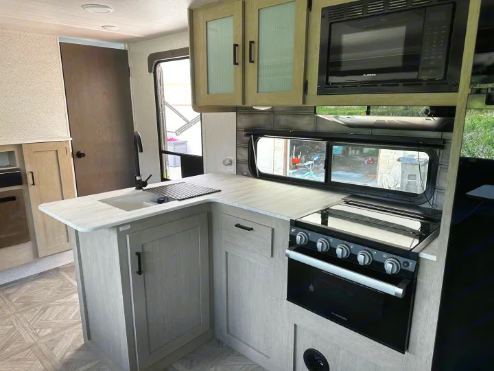 Spacious RV rental kitchen with modern light wood cabinets, light countertop, sink, black microwave, and stove.. Forest River Wildwood X-Lite 2023