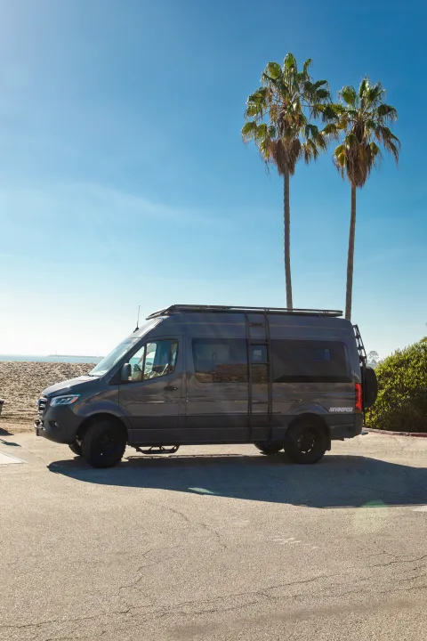 A rugged grey Sprinter camper van, perfect for an rv rental, parked on asphalt by a sunny beach with palm trees.. Mercedes Sprinter 2021