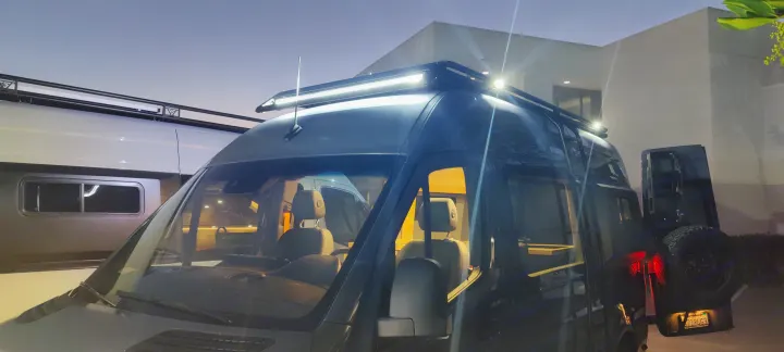 A dark rv rental camper van with bright roof-mounted LED lights shines at dusk, parked outdoors.. Mercedes Sprinter 2021