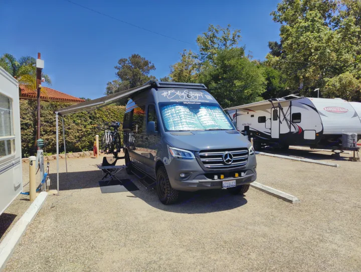 A grey camper van with an extended awning and bikes, parked on gravel alongside an RV. Ideal for your next RV rental trip.. Mercedes Sprinter 2021