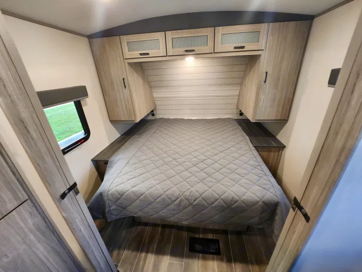 Interior view of an RV rental bedroom showing a bed with a grey comforter, built-in storage, and a window.. Keystone RV Passport GT Ultra Lite 2023