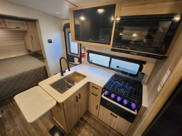 Modern RV rental interior with compact kitchen: sink, stovetop, microwave, TV, and adjacent bed area. Ideal for travel.. Keystone RV Passport GT Ultra Lite 2023